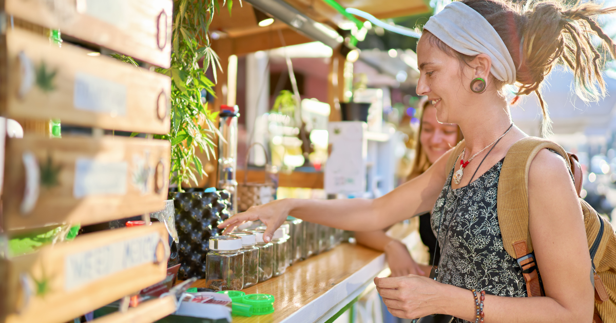 A person is smiling as they examine jars of cannabis products during a 420 celebration. The 420 sales booth is decorated with green plants and wooden signs, creating a natural and inviting atmosphere. 